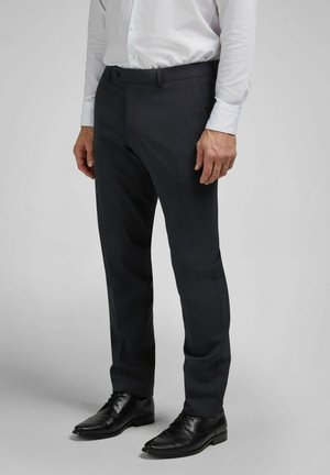 Mann trägt schwarze formelle Hose, schwarze Lederschuhe und ein weißes Hemd vor einem schlichten grauen Hintergrund.