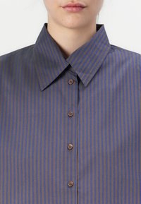 Gros plan sur une personne portant une chemise à boutons rayée verticalement bleu et marron avec un large col.
