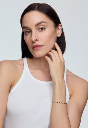 Czarna koralikowa bransoletka na złotym łańcuszku, noszona na jasnej skórze nadgarstka. Osoba nosząca ma proste ciemne włosy i neutralny wyraz twarzy.