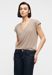 Γυναίκα με κοντά σκούρα μαλλιά, που φοράει μπεζ μπλουζάκι με V-λαιμόκοψη και κοντά μανίκια, καθώς και σκούρο μπλε τζιν, στέκεται μπροστά σε ένα απλό ανοιχτό φόντο.