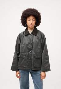 Tamsiai žalia, per didelė striukė su rudu apykakle, dviem priekinėm kišenėm ir mygtukų uždarymu. Audinys atrodo lygus su šiek tiek blizgiu paviršiumi.