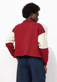 Sweatshirt coloré en rouge et crème, avec un col côtelé et des manches longues, présenté de dos. Tissu doux, ourlet court.