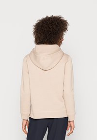Persona con cabello rizado que lleva una sudadera beige y pantalones oscuros, vista desde atrás sobre un fondo blanco liso.
