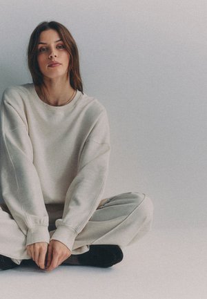 Ung kvinde med brunt hår iført en beige oversized sweatshirt og bukser sidder med krydsede ben på en lys gulv mod en ensfarvet grå baggrund.