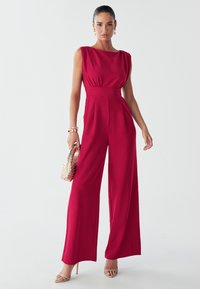 Červený overal s přiléhavým pasem, řasenými rameny a širokými nohavicemi. Hladká látka a čistý střih. Doplňky zahrnují tkanou tašku.