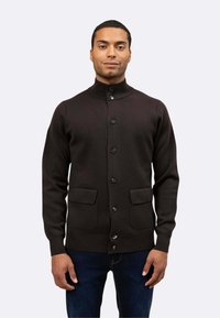 Cardigan marrone scuro realizzato in maglia testurizzata, con colletto alto, bottoni frontali e due tasche anteriori. Indossato sopra jeans blu.