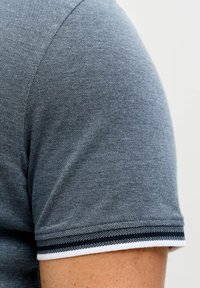 Κοντινό πλάνο του χεριού ενός ατόμου που φοράει μπλε-γκρι πόλο μπλουζάκι με κοντά μανίκια και υφή, με μαύρη και λευκή ριγέ λεπτομέρεια στην άκρη του μανικιού.