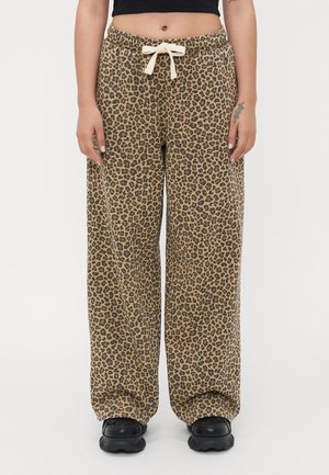 Persoon met losse luipaardprint broek met trekkoord en zwarte schoenen, staand tegen een effen witte achtergrond; het middelste gedeelte is zichtbaar.