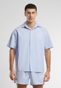 Korte mouwen, knoopshirt in lichtblauw en witte strepen, met een logo op de borst. Gecombineerd met bijpassende shorts, de stof lijkt glad.