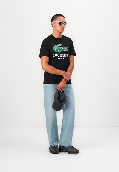 Черна памучна тениска с зелено лого на крокодил и текст "LACOSTE PARIS", комбинирана с светлосини дънки, сива шапка и тъмни обувки.