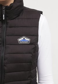Svart pufferväst med dragkedja framtill, horisontell sömnad och ett blått och vitt logotypmärke på bröstet. Texturerat, lätt material.