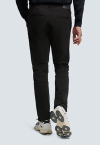 Zwarte katoenen broek met een slim fit, achterzakken, gecombineerd met witte en grijze sportschoenen, weergegeven vanuit de achterkant.