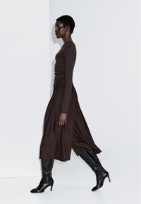 Femme aux cheveux courts portant des lunettes de soleil sombres, un haut à manches longues ajusté, un pantalon large marron foncé et des bottes noires à talons montant jusqu'au genou, marchant de côté.