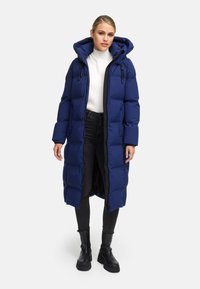 Long navy puffer coat with a hood, featuring zigzag stitching, adjustable toggles, and a front zipper. Worn over a white sweater and black pants.