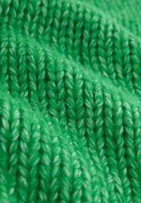 Grønt strikket stof med et struktureret mønster, der har tykke, snoede garntyper og en fuzzy overflade, som viser varierende nuancer af grønt.