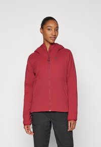 Veste légère rouge avec capuche, dotée d'une fermeture éclair à l'avant et d'un logo discret. Texture lisse et design ajusté, associée à un pantalon sombre.