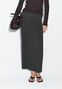 Vrouw draagt een donkere geplooide midirok, een top met lange mouwen, zwarte sandalen en houdt een losse donkere leren handtas naast zich vast.