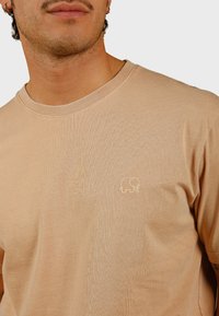 Licht beige T-shirt van zacht materiaal, met een ronde hals en korte mouwen. Bevat een klein geborduurd olifantenlogo op de borst.