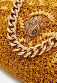 Zlatá tkaná kabelka s texturovaným povrchem, vybavená metalickým řetízkovým popruhem a dekorativním zapínáním s rhinestone kamínky a akcentem s drahokamem.