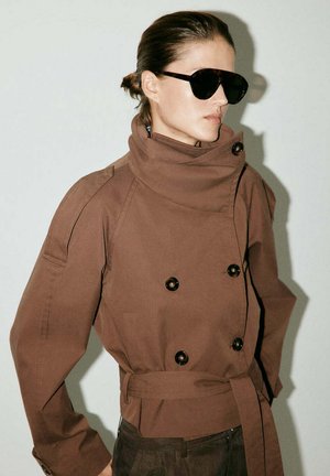 Mujer que lleva una chaqueta marrón de doble botonadura extragrande con cuello alto y cinturón, gafas de sol oscuras, cabello recogido, mirando hacia la derecha sobre un fondo liso.