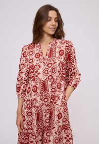 Young woman in a knee-length, loose-fitting dress with red floral and geometric patterns, three-quarter sleeves, and hands relaxed.