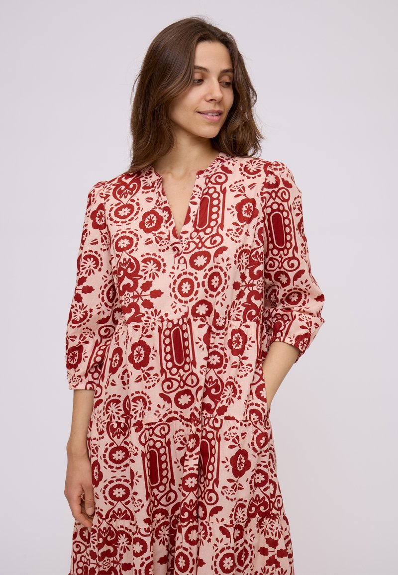 Young woman in a knee-length, loose-fitting dress with red floral and geometric patterns, three-quarter sleeves, and hands relaxed.
