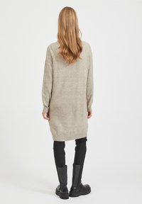 Long pull beige avec ourlet et poignets côtelés, texture tricotée, associé à un pantalon noir ajusté et des bottes noires montantes jusqu'aux genoux.