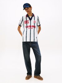 Weißes Poloshirt mit blauen vertikalen Streifen, roter "85" und "Tommy Jeans"-Schriftzug, kombiniert mit dunklen Jeans und braunen Sneakers. Marineblaue Mütze.