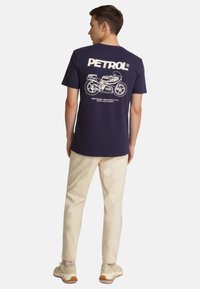 Camiseta de algodón azul marino con estampado gráfico blanco que presenta una motocicleta y el texto "PETROL" en la parte posterior. Combinada con pantalones beiges y zapatillas.