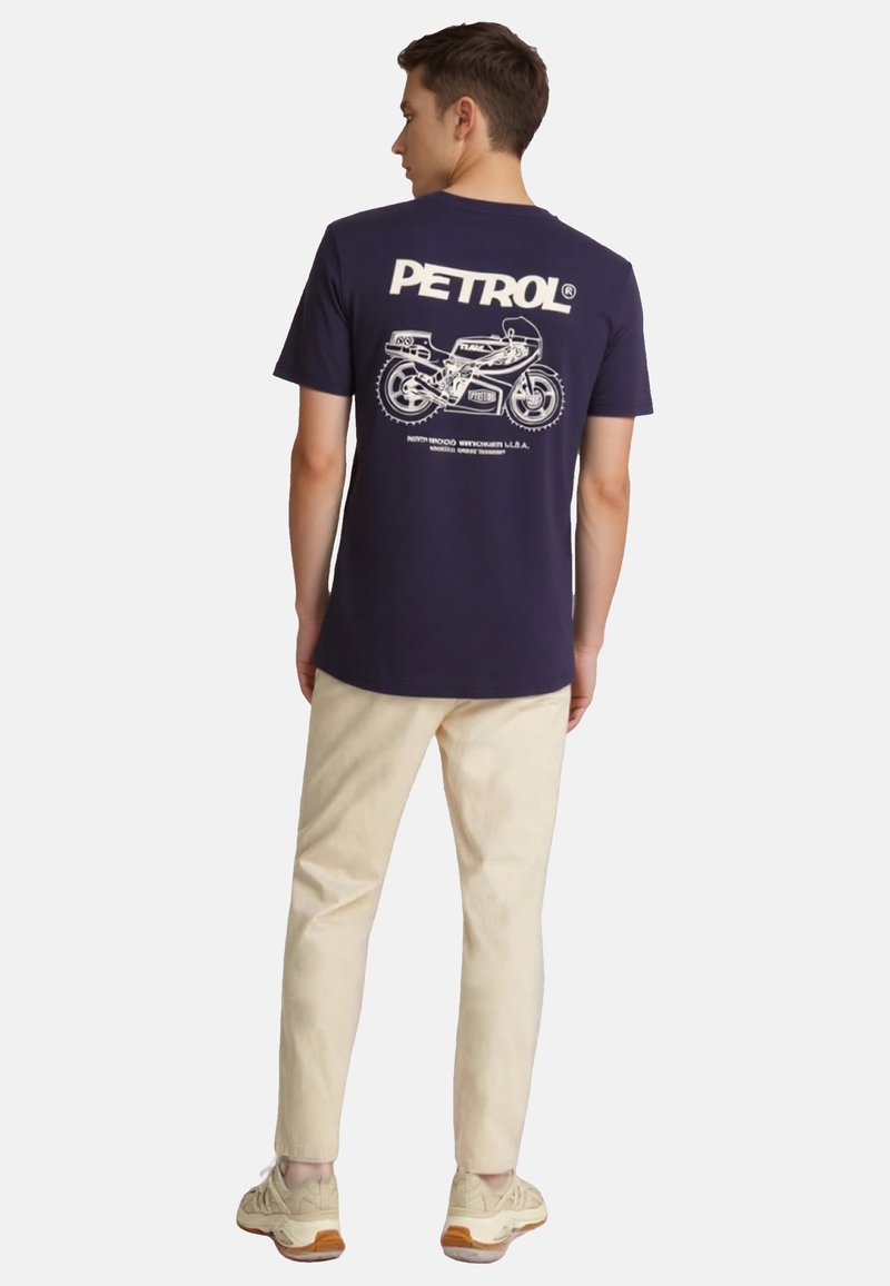 Camiseta de algodón azul marino con estampado gráfico blanco que presenta una motocicleta y el texto "PETROL" en la parte posterior. Combinada con pantalones beiges y zapatillas.