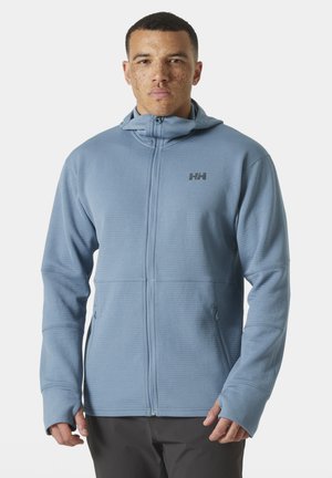 Man wearing a light blue zippered fleece jacket with hood and thumb holes, standing against a plain background.