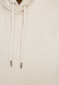 Felpa leggera beige con cappuccio regolabile, caratterizzata da un tessuto testurizzato, estremità metalliche sui cordini e un piccolo logo discreto sul petto.