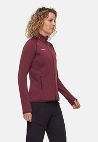 Giacca softshell bordeaux con colletto alto, zip frontale completa e due tasche laterali. Presenta un design aderente e una texture liscia.