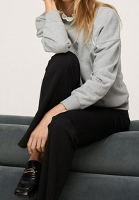 Szary sweatshirt z okrągłym dekoltem i ściągaczami, w połączeniu z czarnymi spodniami. Czarne mokasyny z złotym akcentem. Siedząca postawa na miękkiej powierzchni.