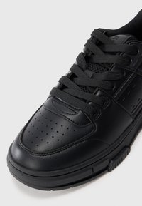 Black sneaker with textured leather upper, layered design, perforated toe box, flat black laces, and rubber outsole with traction grooves.