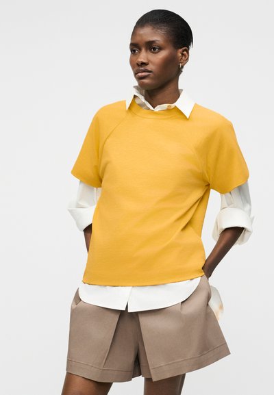 Femme aux cheveux courts, portant un haut jaune à manches courtes superposé à une chemise blanche et un short beige arrivant aux genoux, debout les mains dans les poches.