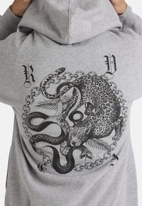 Grijze hoodie met zwart grafisch ontwerp van een leeuw en een slang samengeslingerd binnen een cirkelvormig kettingpatroon. Met capuchon en lange mouwen.