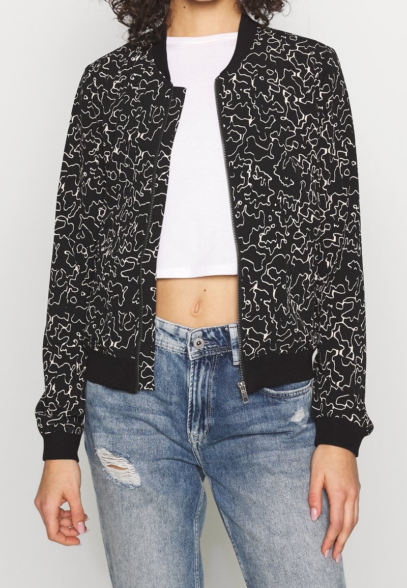 Blouson aviateur noir avec un motif abstrait blanc, poignets et ourlet côtelés, et une fermeture éclair à l'avant. Porté sur un haut court blanc.