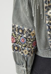 Denimjakke-erm med blomsterbroderi og paljett-detaljer i blått, gull, rødt og krem på svarte og beige stoffbånd.