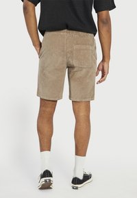Homme portant un short en velours côtelé beige, une chemise noire à manches courtes avec texture, des chaussettes blanches et des baskets noires à tige basse, se tenant de dos.