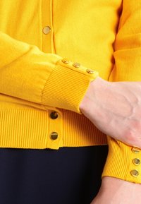 Gul ribbad kofta med guldfärgade knappar vid ärmslut och fram, med en mjuk textur och figurnära design.