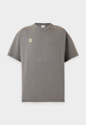 Άνδρας με κοντά μαλλιά φοράει άνετο γκρι t-shirt με κίτρινη γοτθική γραμματοσειρά "Arket Copenhagen" στην πλάτη, σε λευκό φόντο.