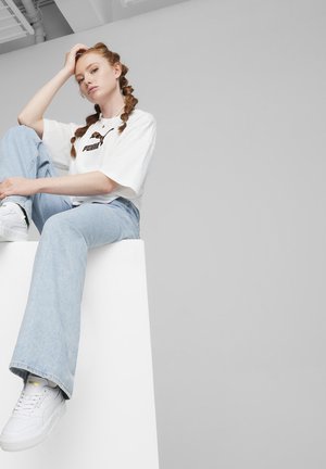 T-shirt blanc surdimensionné avec un logo noir, jean large bleu clair, et baskets blanches avec des accents dorés, assis sur une plateforme blanche.