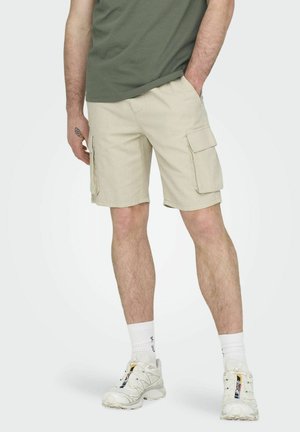 Beige Cargo-Shorts mit zwei seitlichen Taschen, aus leichtem Stoff gefertigt. Getragen mit einem grünen T-Shirt und weißen Sneakers.