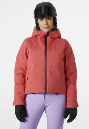 Rode, dikke skijas met een ritssluiting aan de voorkant, verstelbare capuchon en zijzakken, gecombineerd met paarse skibroek en zwarte handschoenen.