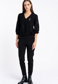 Kobieta ubrana w czarną, strukturalną bluzkę z dekoltem w kształcie litery V i rękawami do trzech czwarte, czarne dopasowane spodnie oraz czarne botki na kostkę, stojąca na białym tle.