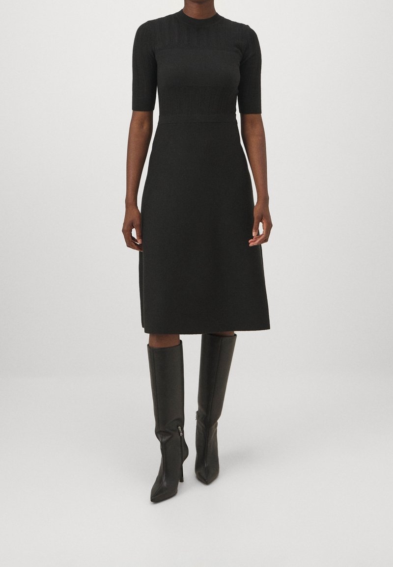 Femme portant une robe noire mi-longue à manches courtes et des bottes noires à talons montant jusqu'aux genoux, debout devant un fond uni.