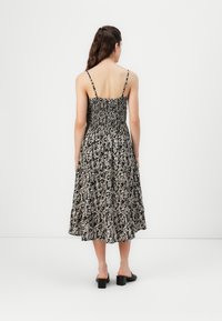 Femme aux longs cheveux portant une robe midi florale noire et blanche à fines bretelles et des chaussures noires à petit talon carré, debout contre un mur blanc uni.