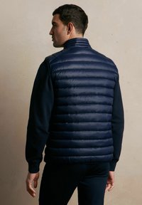 Granatowa kamizelka puchowa z poziomymi szwami, wysokim kołnierzem i gładką teksturą, noszona na czarnym topie z długim rękawem.