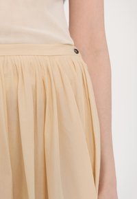 Jasno beżowa spódnica z plisami, wykonana z teksturowanej tkaniny, z pasem w talii i czarnym guzikiem. Slim fit, o długości midi.